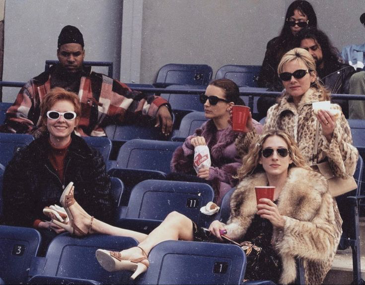 Carrie, Miranda, Charlotte y Samantha con abrigos de piel y gafas de sol en las gradas de un estadio